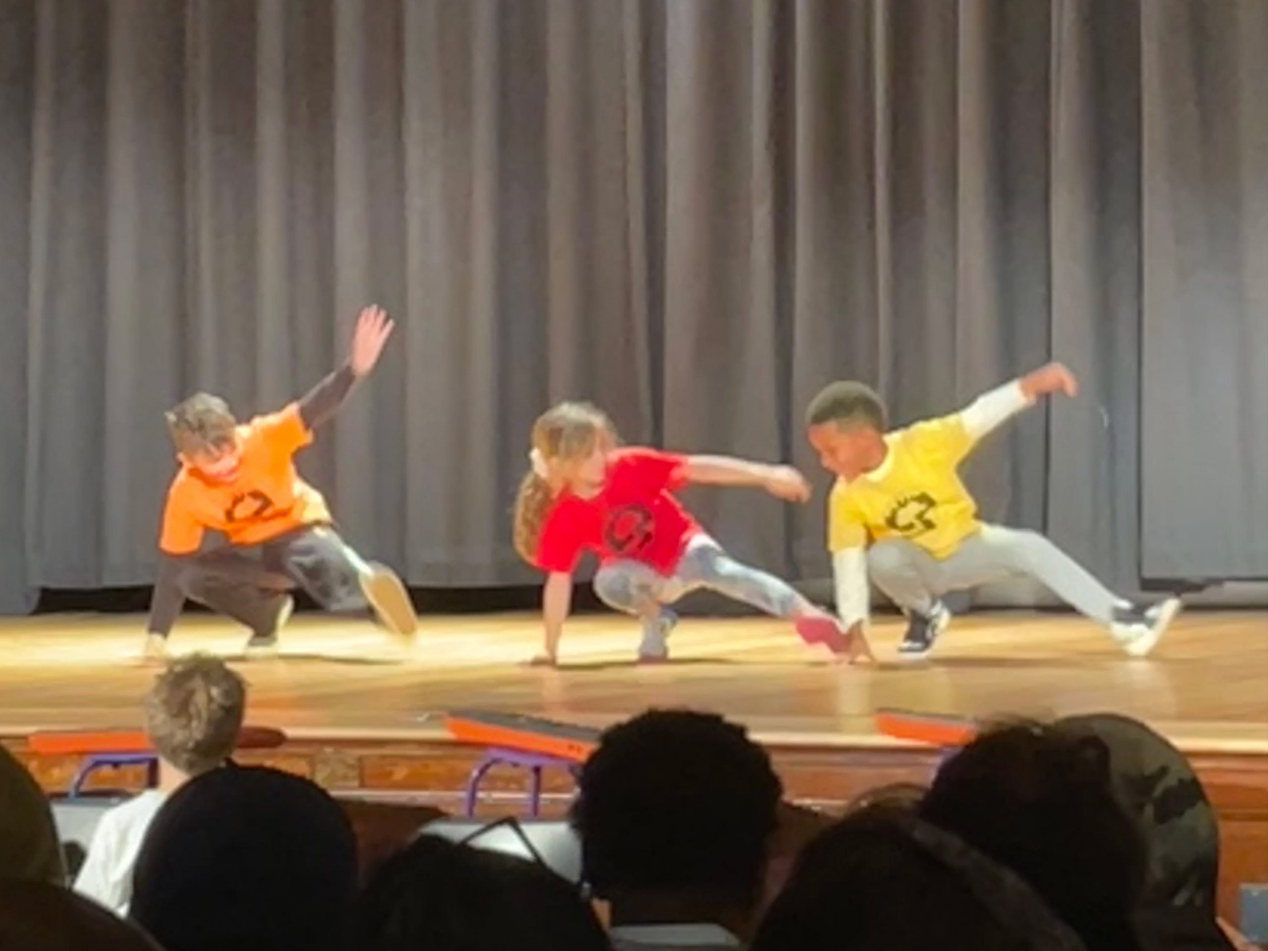 three young students each in orange, red, and yellow t-shirts dance on a stage in front of an audience of family and community members